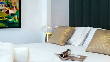 Una cama bien hecha con almohadas blancas y doradas, una revista y una lámpara de vidrio sobre una superficie blanca.