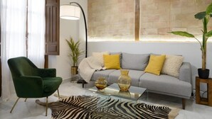 A living room with a green armchair, a grey sofa with yellow cushions, a zebra-patterned rug, and a large window with a stone wall. - Apartment Riad Alhambra 3 (granada)