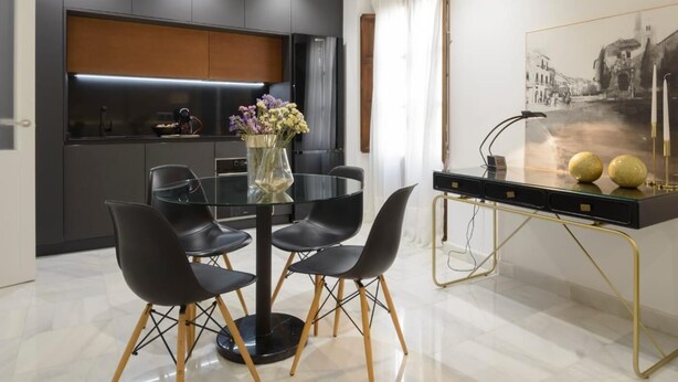 A modern dining area with a glass table surrounded by black chairs, a vase with flowers, and a black and gold side table with decorative spheres. - Apartment Riad Alhambra 3 (granada)