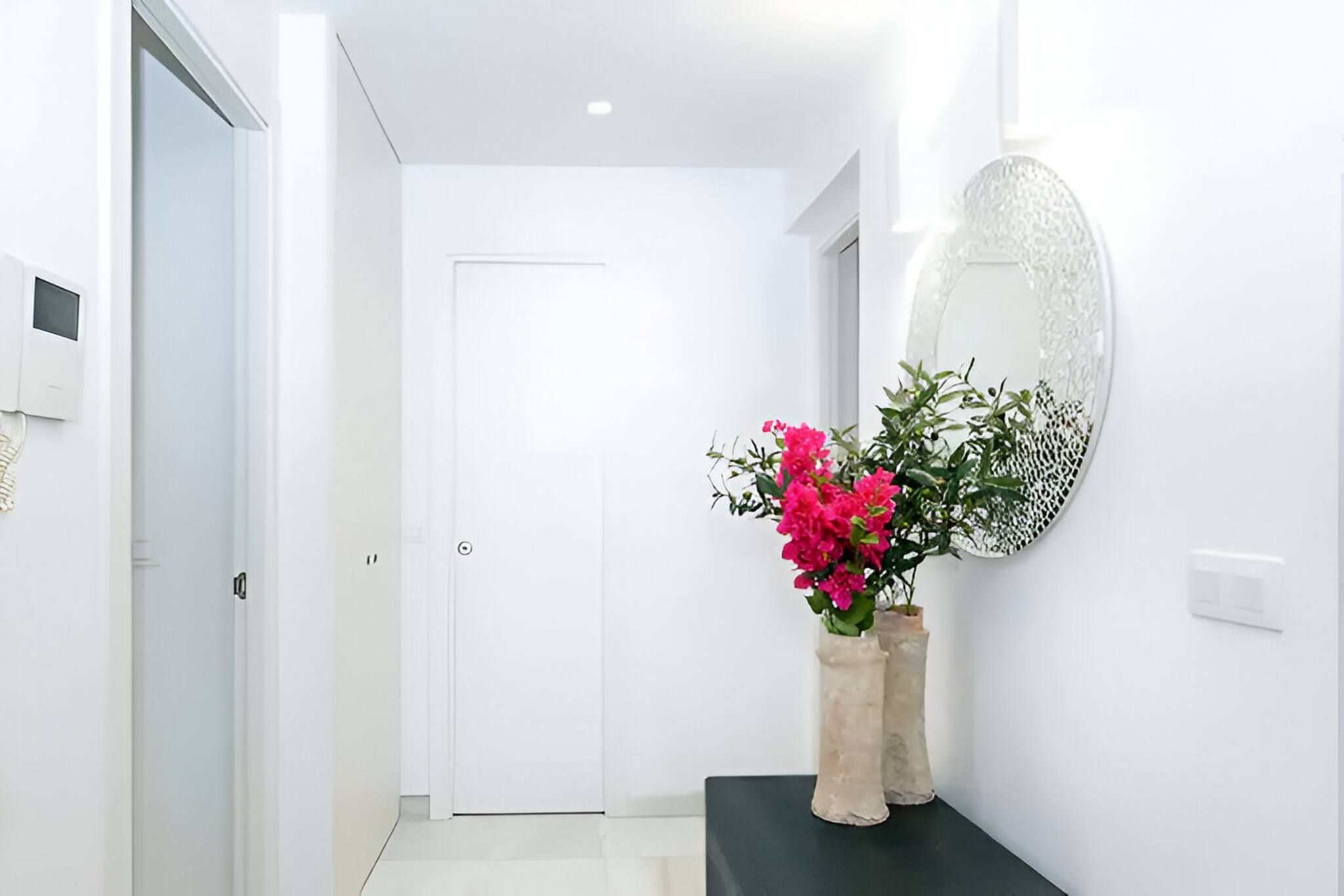 Un pasillo con una puerta blanca, una consola negra y un jarrón de flores rosadas, además de un espejo decorativo en la pared.