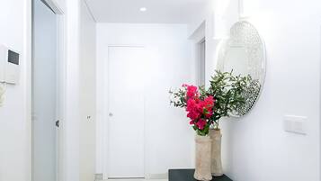 Un pasillo con una puerta blanca, una consola negra y un jarrón de flores rosadas, además de un espejo decorativo en la pared.