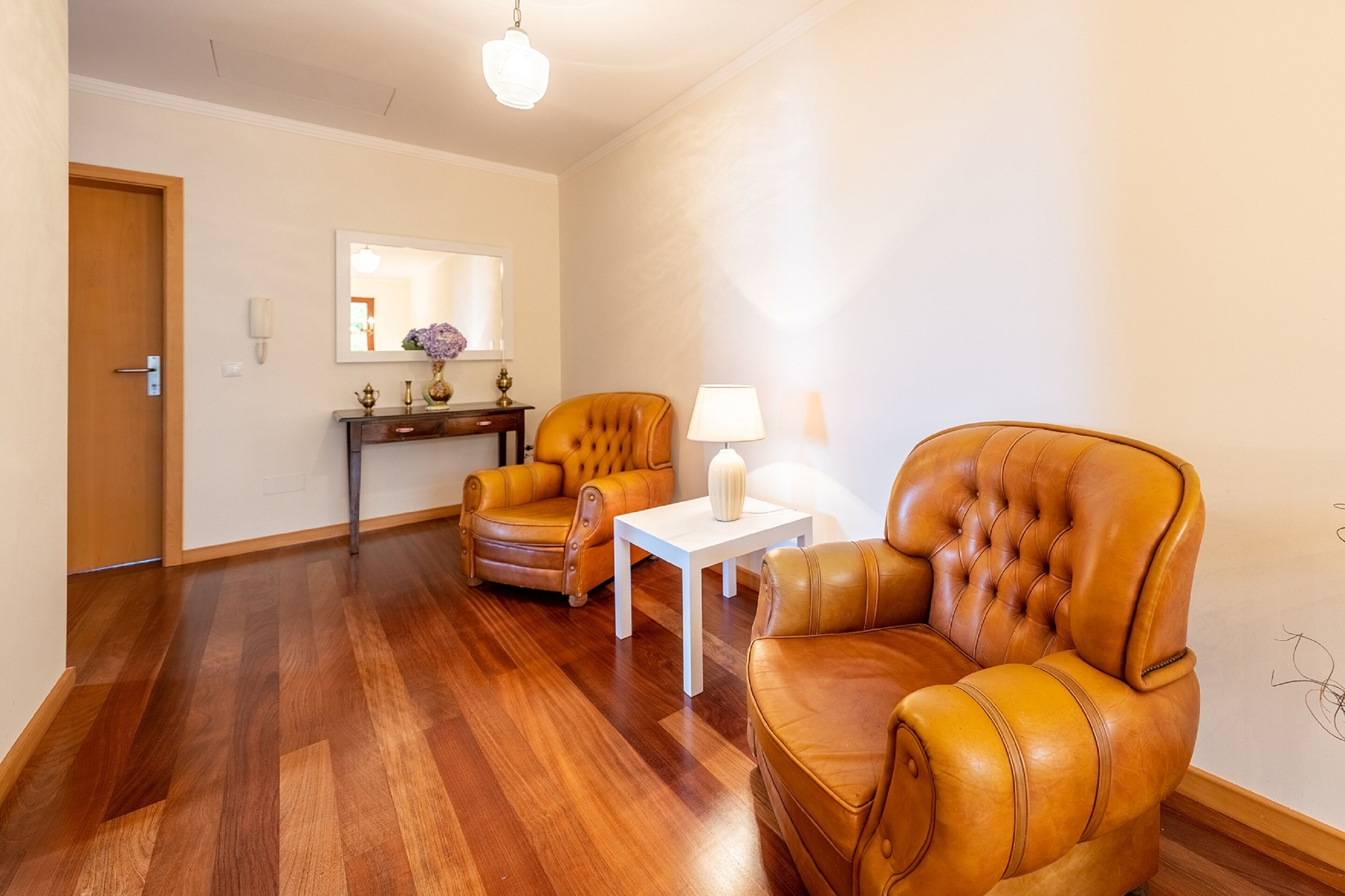 A spacious room with a wooden floor, two leather armchairs, a white table, and a mirror above a console table with decorative items.