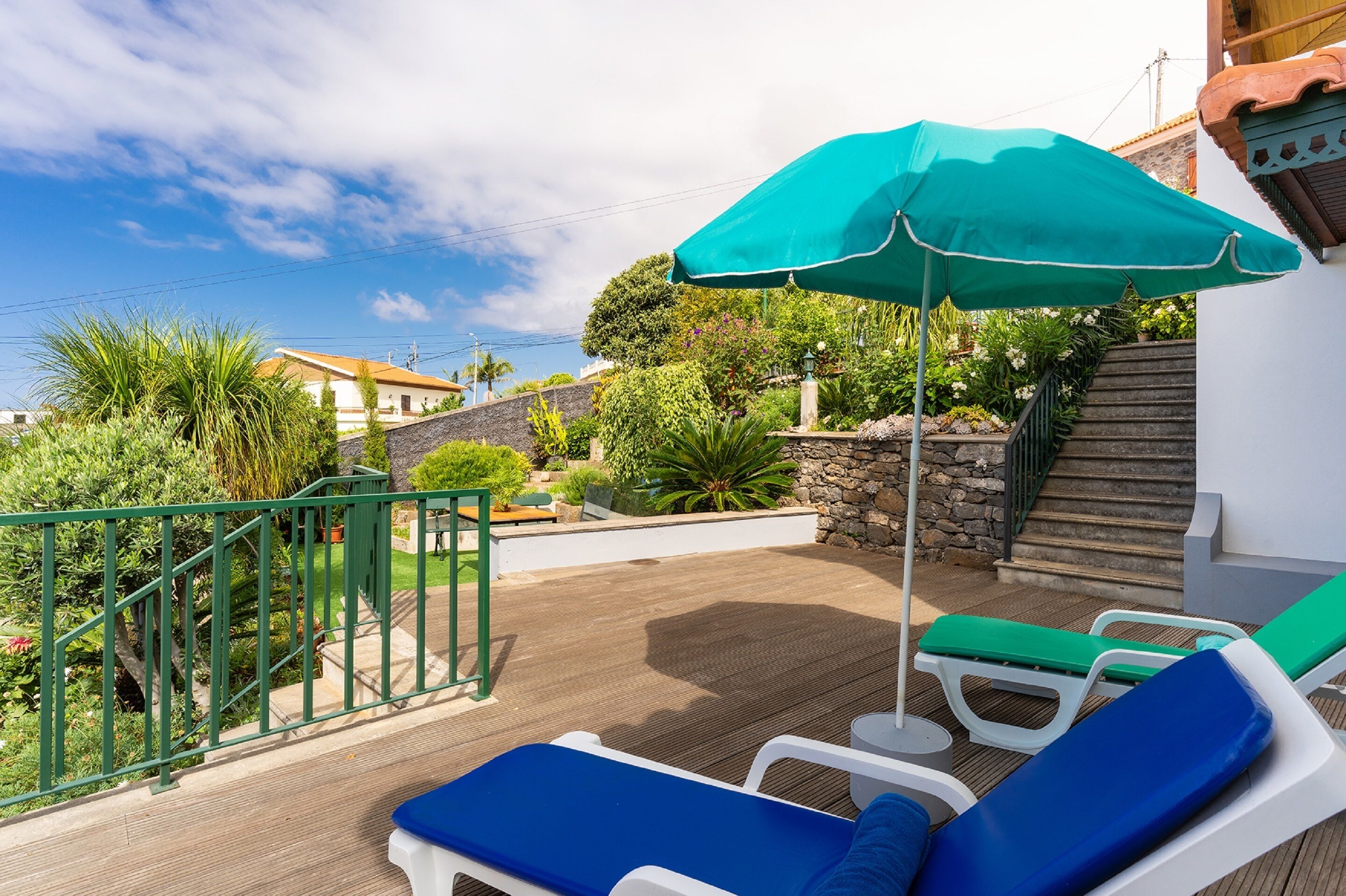 Een houten terras met lounge fauteuils en een groene parasol, omgeven door groen en een stenen muur.