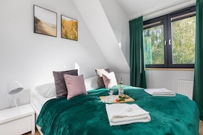 Une chambre à coucher avec un lit, des oreillers, une table de chevet, une lampe, une fenêtre avec des rideaux et deux tableaux encadrés accrochés au mur.