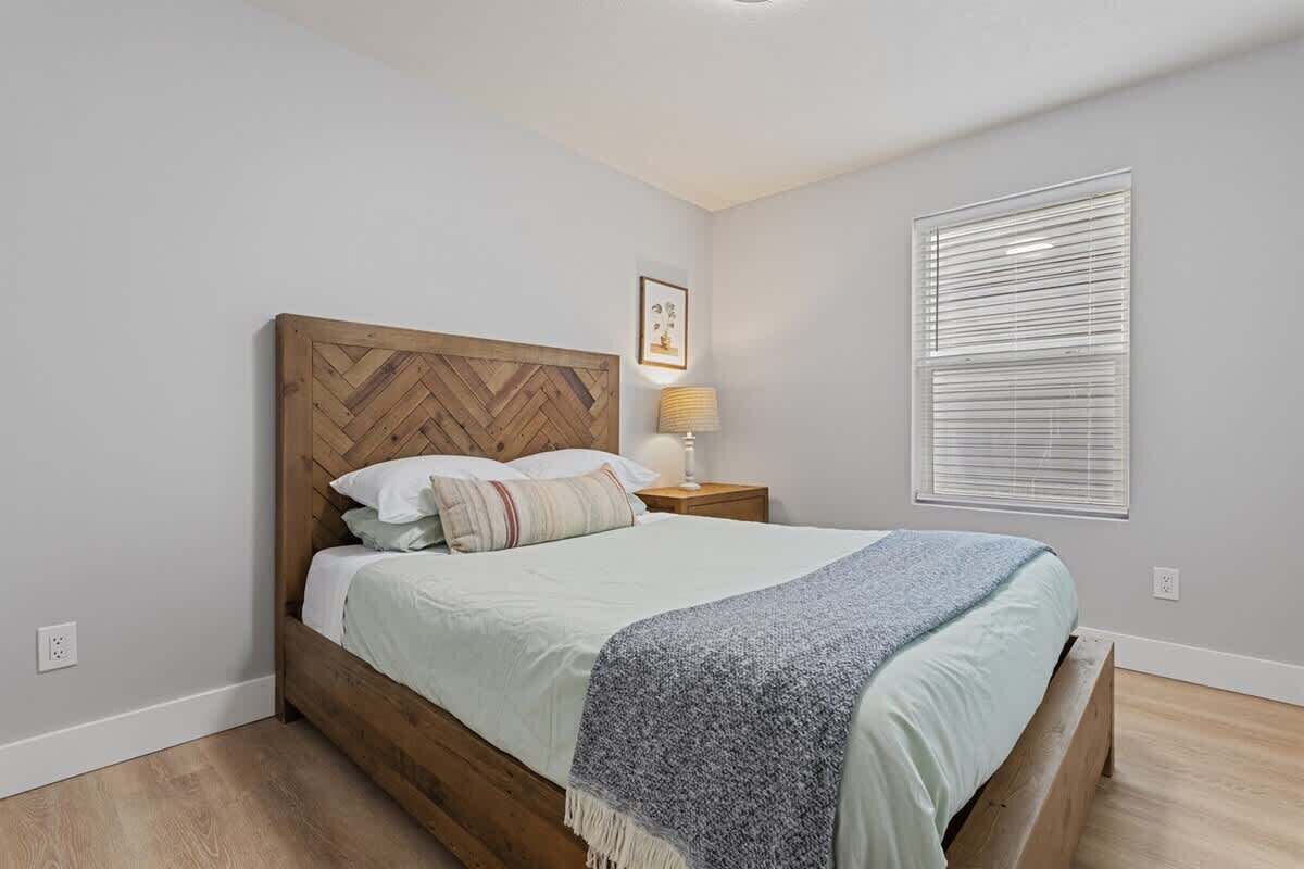Un dormitorio con cabecera de madera, cama con cubrecama de color claro y manta azul, lámpara de noche, cuadro enmarcado en la pared y ventana con persianas.