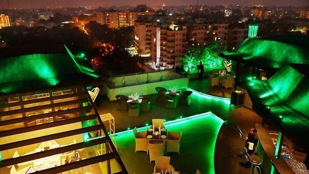 Un bar en plein air situé sur un toit, doté d’un éclairage d’ambiance verdâtre, d’un aménagement avec des espaces pour s’asseoir et offrant une vue sur le paysage urbain la nuit.