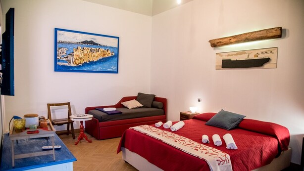 A hotel room with a bed, a red sofa, a small table, a painting, and a wooden shelf.