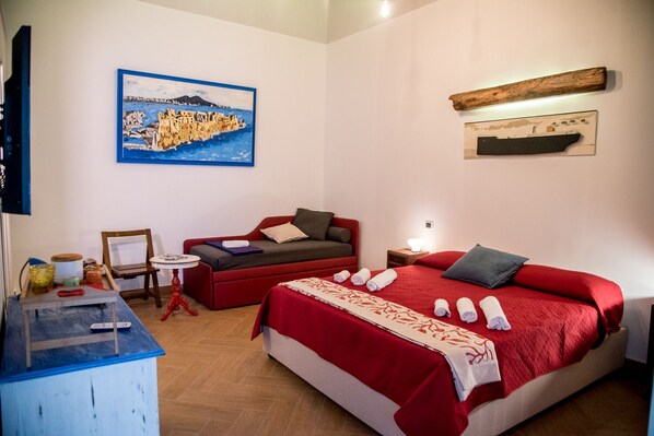 A hotel room with a bed, a red sofa, a small table, a painting, and a wooden shelf.