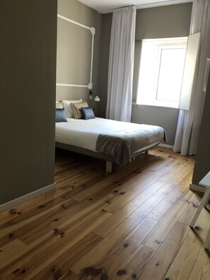 A bedroom with a bed, wooden flooring, a window with curtains, and a nightstand.