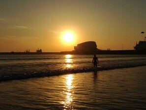 A sunset over a body of water with a large ship silhouette and a person standing in the water.