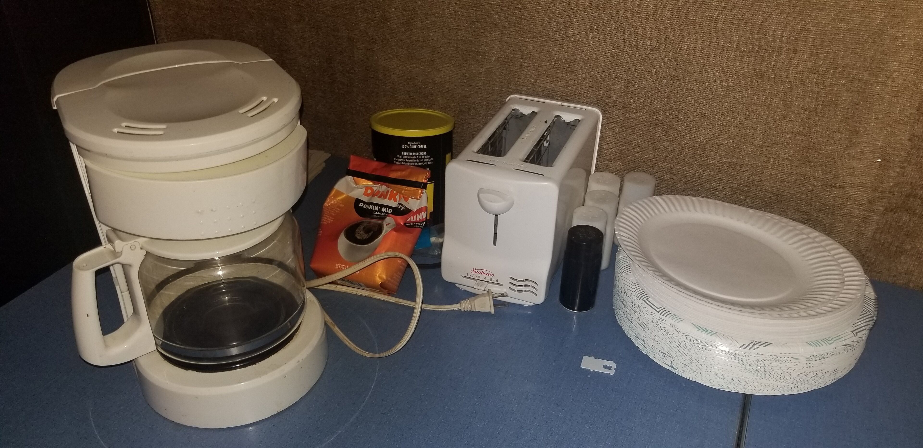 Ett litet kök med en kaffebryggare, brödrost, papperstallrikar och en burk kaffe.