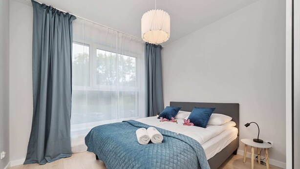 A bedroom with a bed, a bedside table, a window with curtains, and a hanging light fixture.