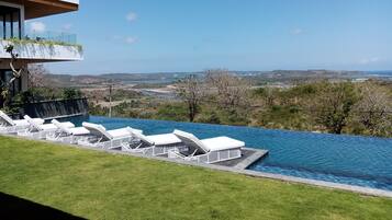 Une piscine à débordement bordée de chaises longues blanches, une vue dégagée sur le paysage et un bâtiment moderne doté d’un balcon.