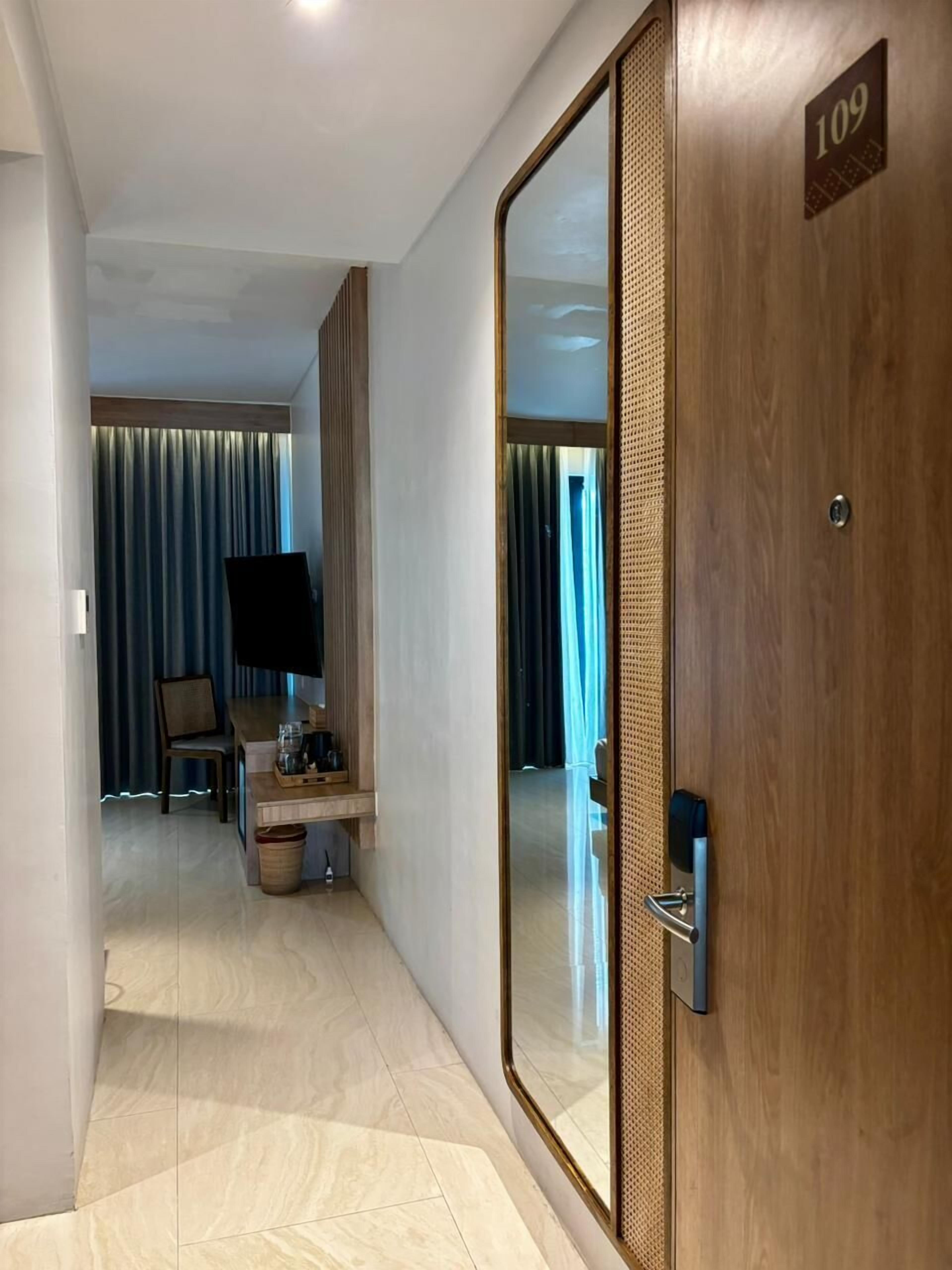 Ett hotellrum med en stor spegel, en trädörr med ett nummer på, ett litet bord med en kaffebryggare, en stol och en tv.