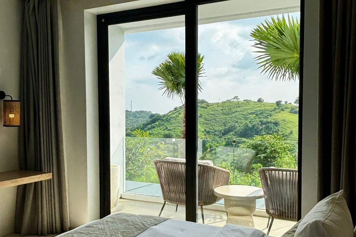 Quarto de hotel com cama, uma mesinha e vista para uma paisagem exuberante através de uma grande janela.