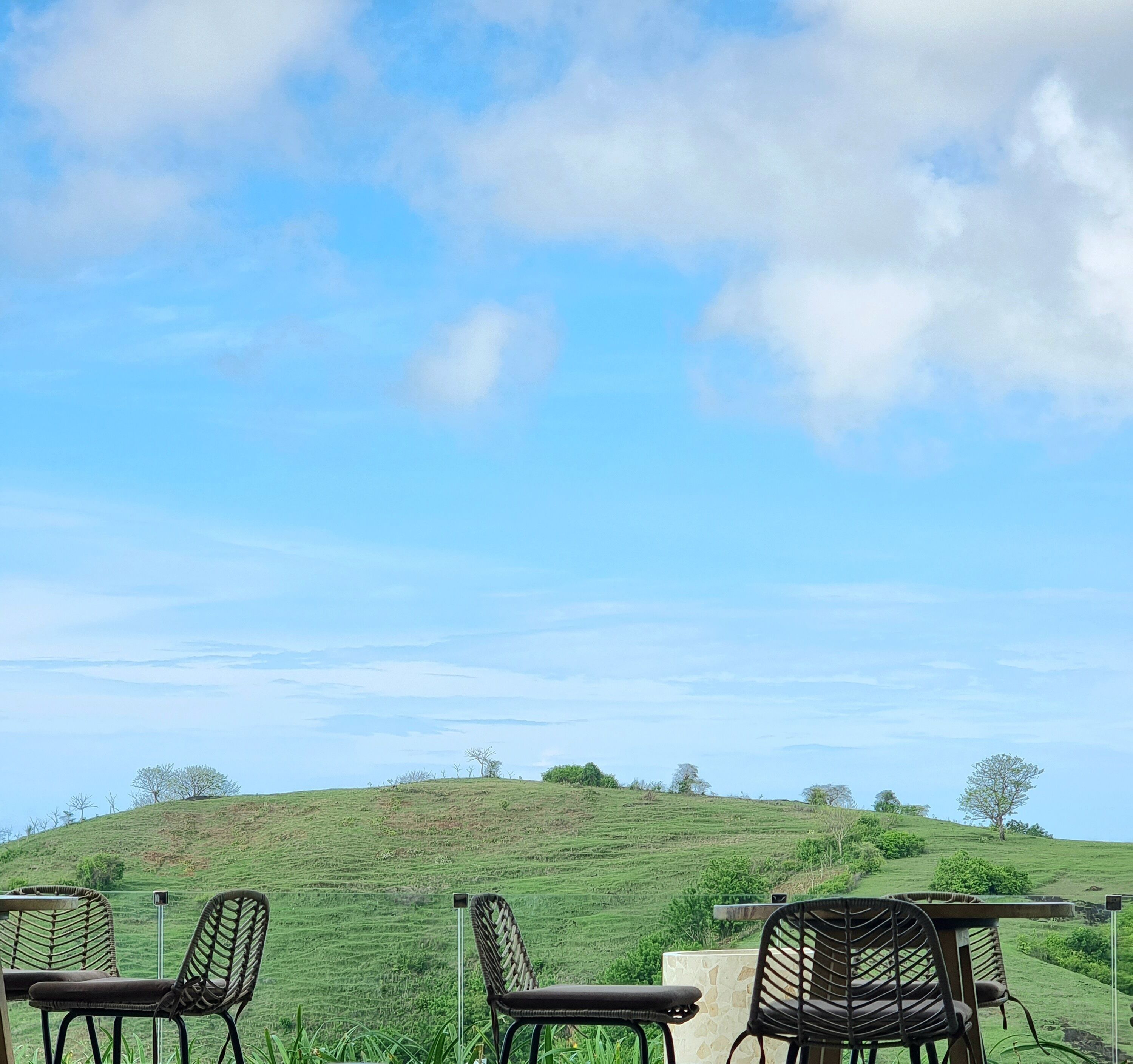 A terrace with wicker chairs and a table, overlooking a green hillside with sparse trees under a clear sky.