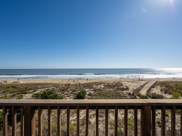 Une vue sur la plage depuis une terrasse en bois, avec un ciel dégagé et l’océan en arrière-plan.