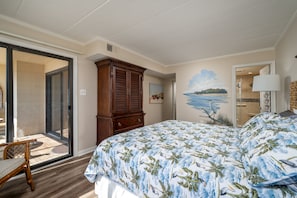 Un dormitorio con una cama grande, un armario de madera, una puerta corrediza de vidrio que da a un balcón y un mural de playa en la pared.