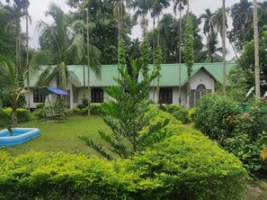 A bungalow with a green roof, surrounded by lush greenery and palm trees.