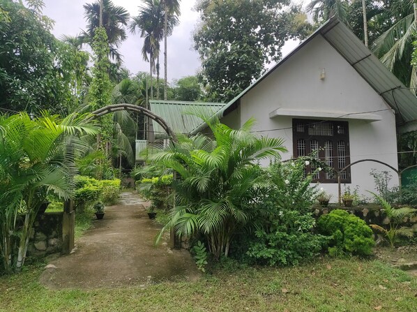 A white house with a green roof, surrounded by lush greenery and a narrow pathway.
