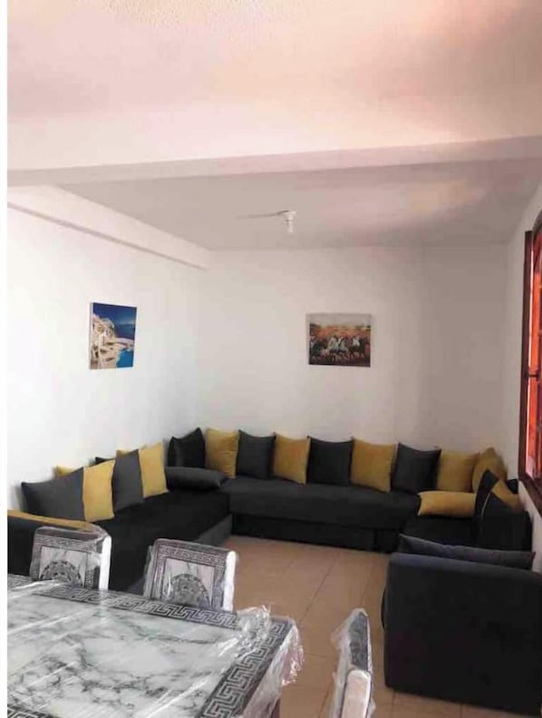 Un salon avec un canapé foncé, des coussins jaunes, une table basse en marbre et deux tableaux accrochés au mur.