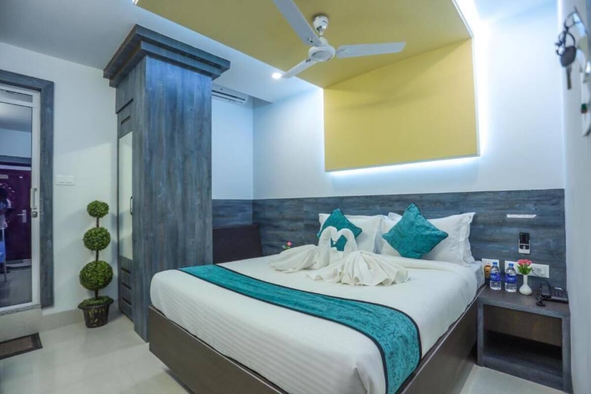 Ein modernes Hotelzimmer mit Bett, Deckenventilator, kleiner Topfpflanze und Nachttisch mit Telefon und Wasserflaschen.