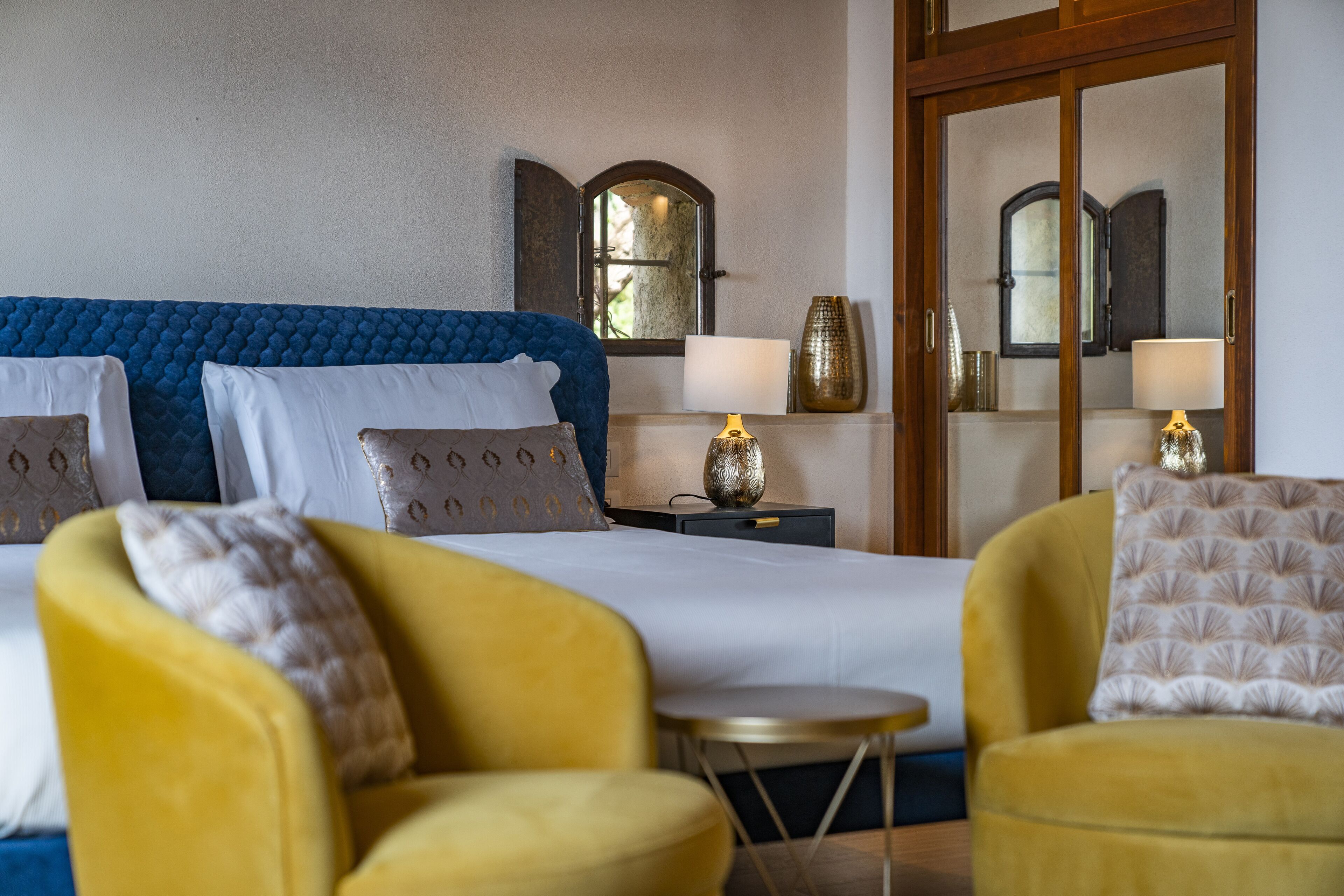 Een hotelkamer met een groot bed, twee gele fauteuils, een klein rond tafeltje en twee bedlampjes.