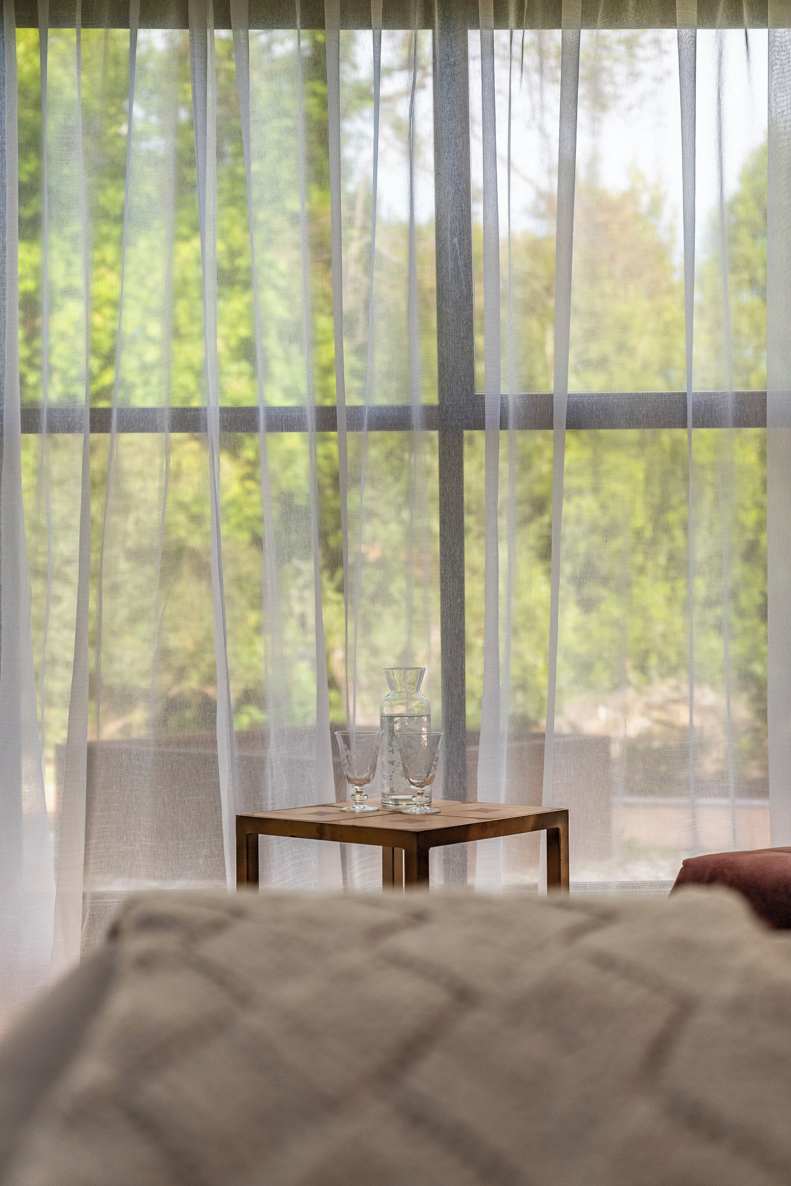 Une chambre avec un lit, une table sur laquelle sont posés deux verres, et une vue sur la verdure à travers des rideaux transparents.