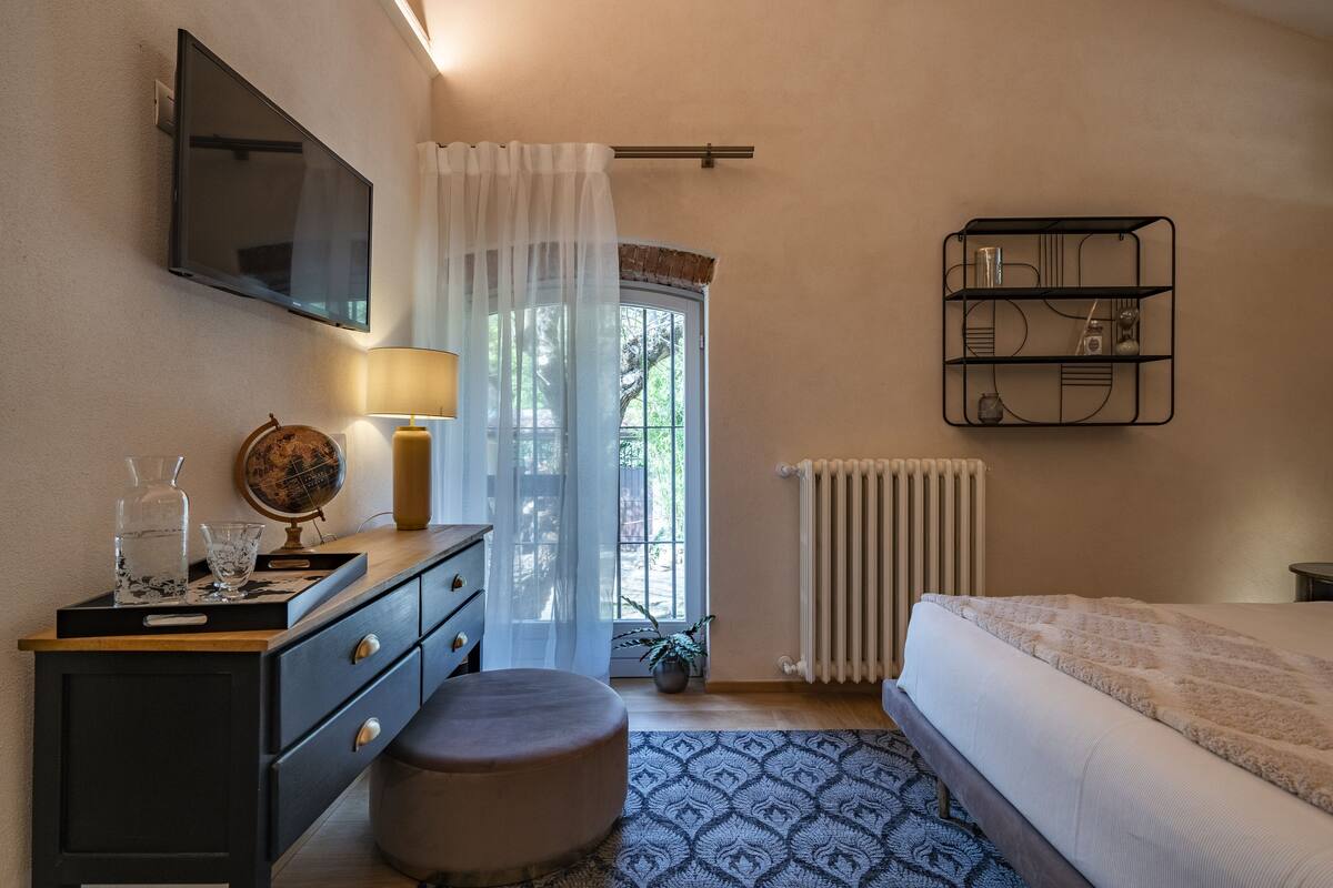 A bedroom with a bed, a dresser, a TV, a window with curtains, a radiator, and a shelf with items.