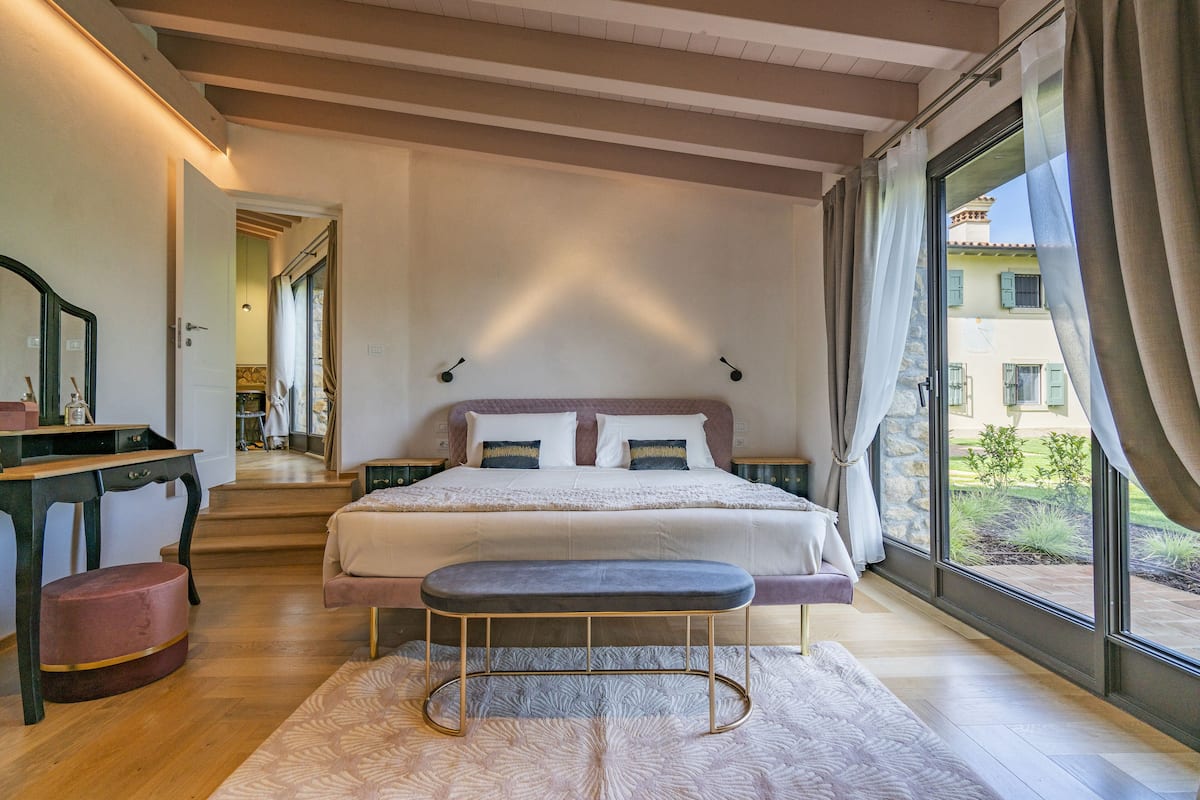 A bedroom with a large bed, a desk, and a bench. There is a window with curtains and a view of the outside.