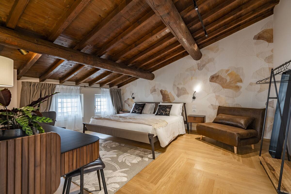 A bedroom with a wooden ceiling, a bed, a sofa, a desk, and a plant.
