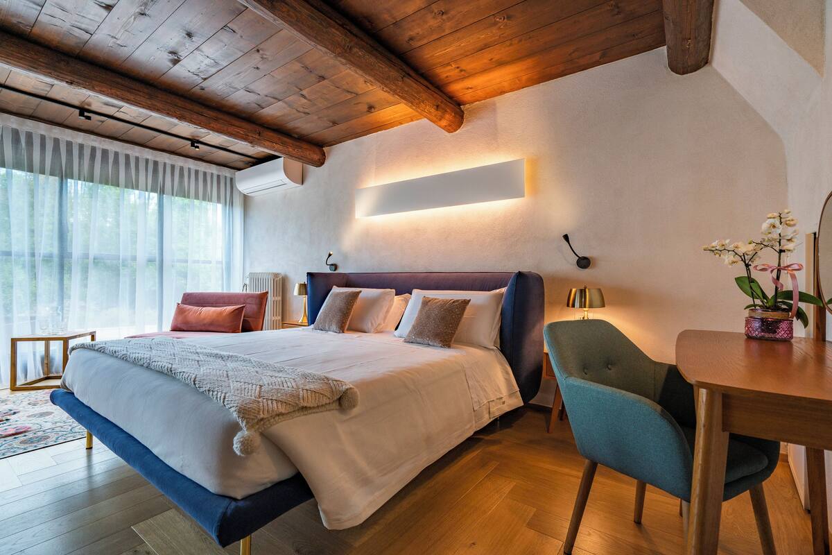 A bedroom with a large bed, wooden ceiling beams, a bedside table with a lamp, and a view of greenery outside.