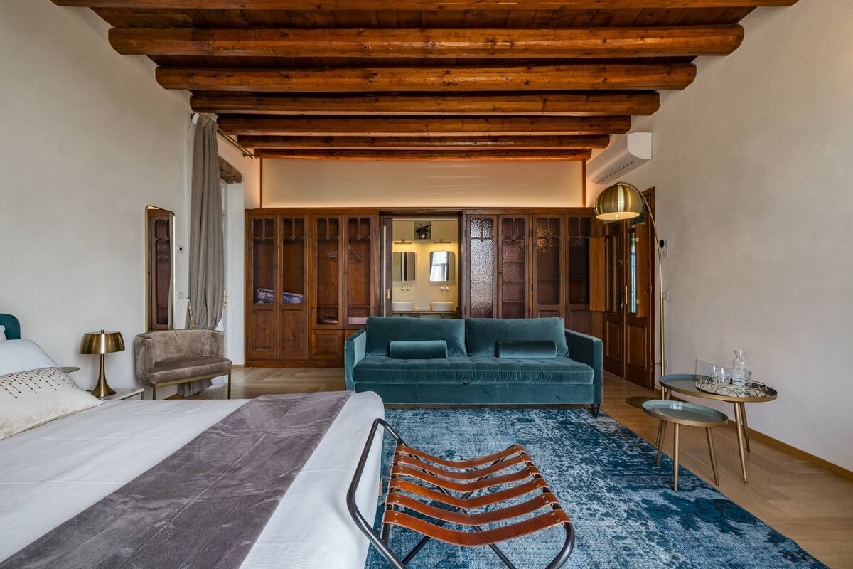 A spacious bedroom with a large bed, a blue sofa, a wooden ceiling, and a small round table with a lamp.