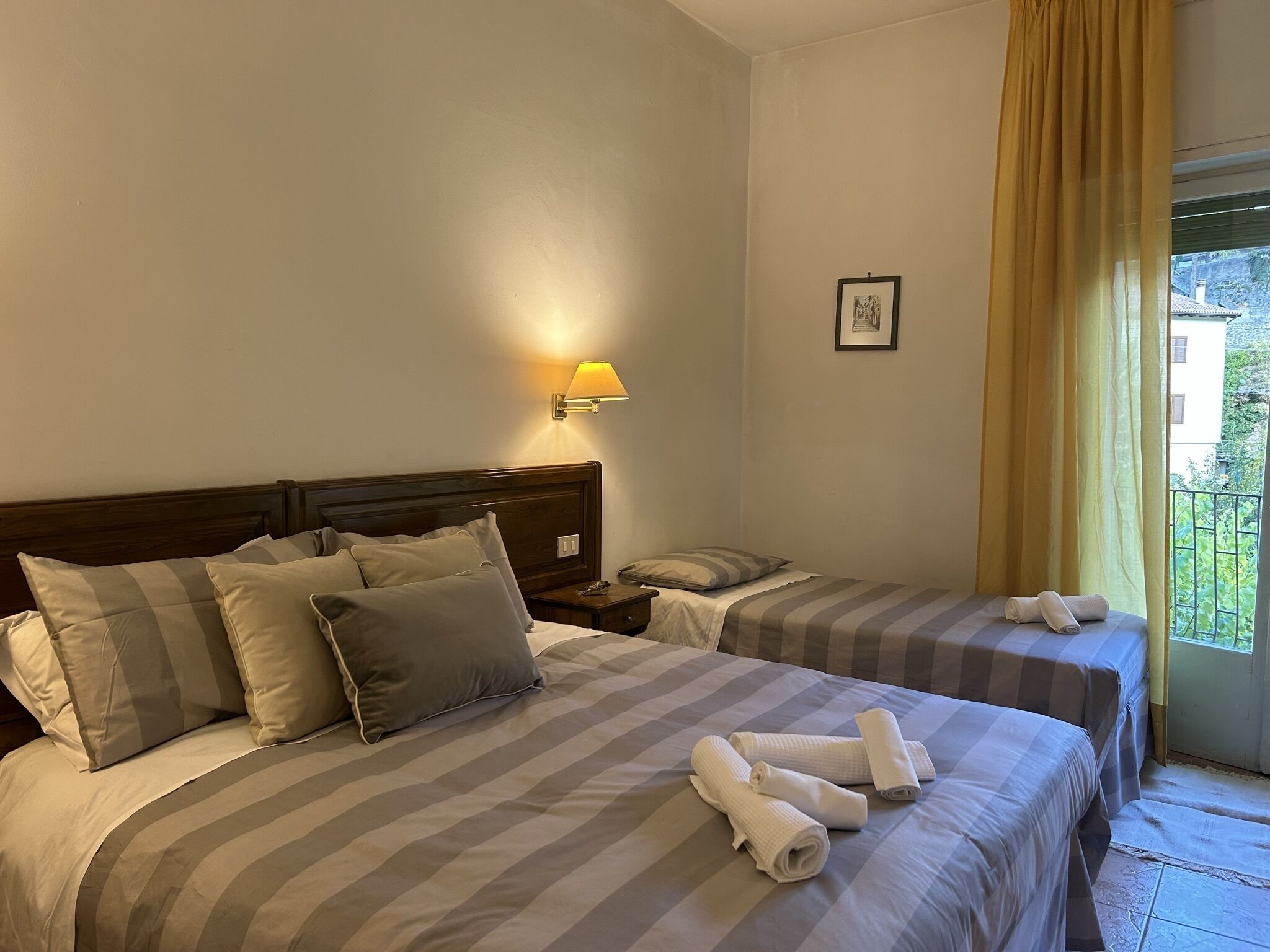 A hotel room with two beds, a nightstand, a lamp, a framed picture, and a view of greenery outside.