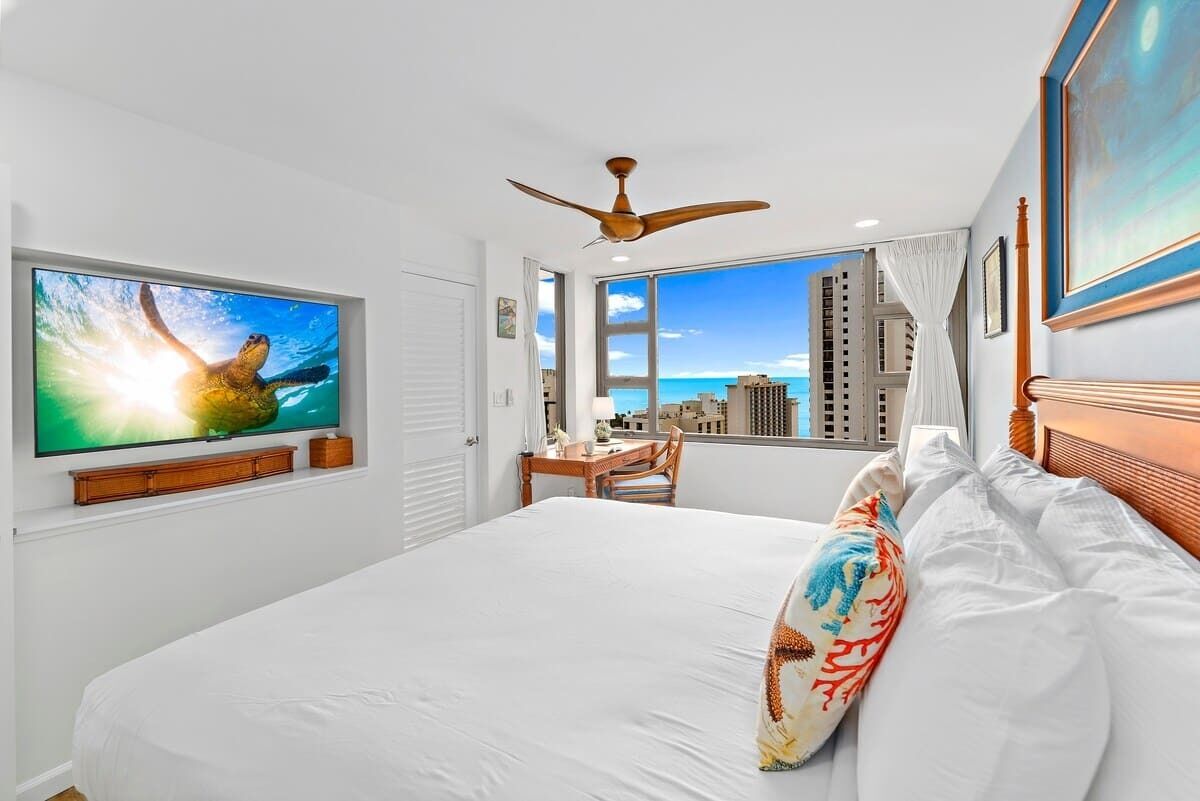 Camera d'albergo con un letto grande, una ventola a soffitto, un televisore che mostra una tartaruga marina, una scrivania con una sedia e vista sulla città attraverso ampie finestre.