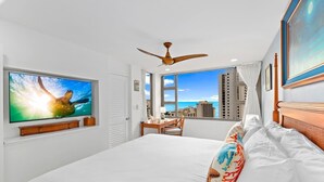 Ein Hotelzimmer mit einem großen Bett, einem Deckenventilator, einem Fernseher, der eine Meeresschildkröte zeigt, einem Schreibtisch mit Stuhl und Blick auf die Stadt durch große Fenster.