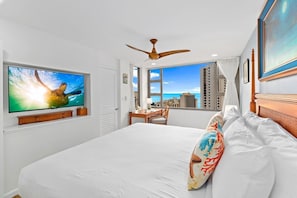 Quarto de hotel com cama grande, ventilador de teto, televisão exibindo uma tartaruga marinha, escrivaninha com cadeira e vista da cidade através de grandes janelas.