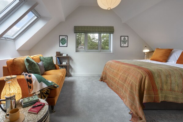 A cozy bedroom with a sloped ceiling, a large window with a view of greenery, a bed with an orange blanket, a bedside table with a lamp, and a reading chair with a lamp. - Barakat Lakes Escape (Ambleside)