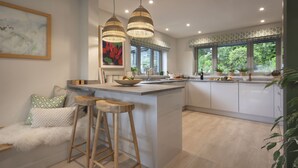 A modern kitchen with a central island, wooden stools, pendant lights, and large windows with a view of greenery. - Barakat Lakes Escape (Ambleside)