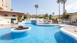 A swimming pool with a circular rock feature, surrounded by palm trees and a multi-story building with balconies.