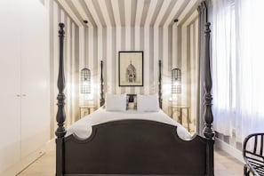 A bed with a dark headboard, white bedding, and a framed picture above it. There are two bedside tables with lamps, and the room has striped walls.