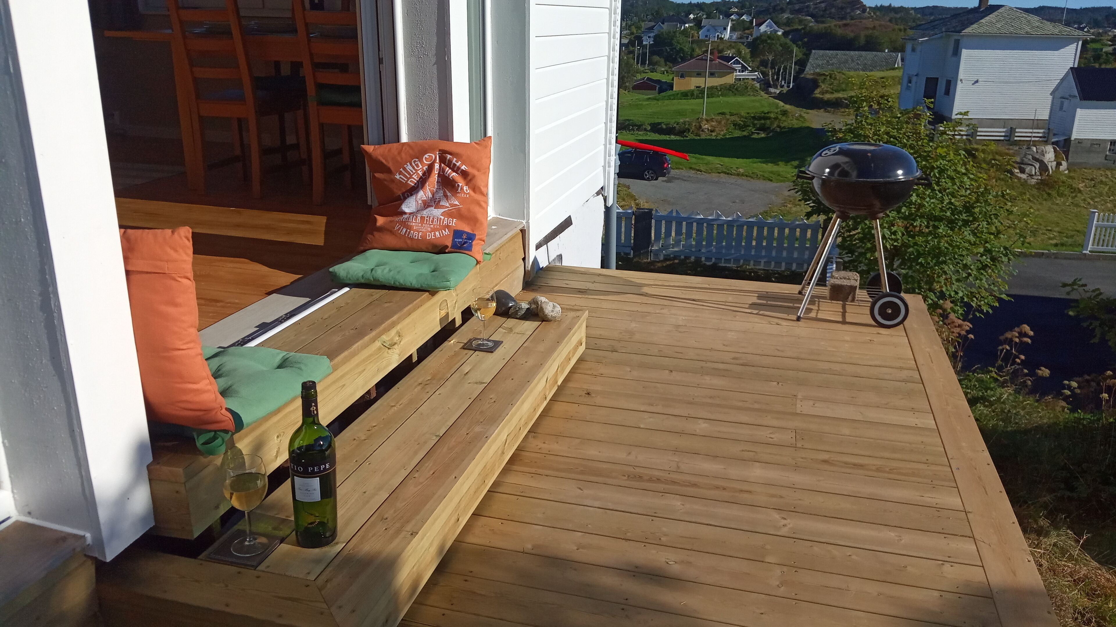 A wooden deck with a bench, a bottle of wine, and a glass on the left, and a barbecue grill on the right.