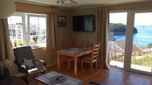 Un salón amplio con una mesa de comedor de madera, asientos cómodos, un televisor de pantalla plana y ventanas grandes que ofrecen una vista panorámica del mar y las colinas.