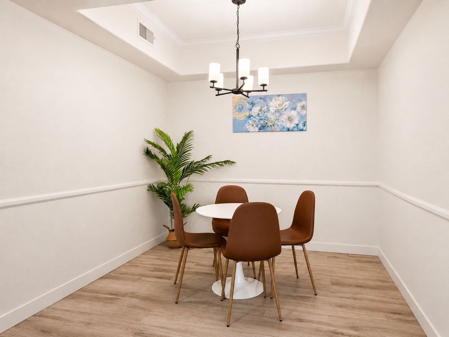 Un área de comedor con una mesa redonda, cuatro sillas marrones, una planta en maceta y un cuadro en la pared.