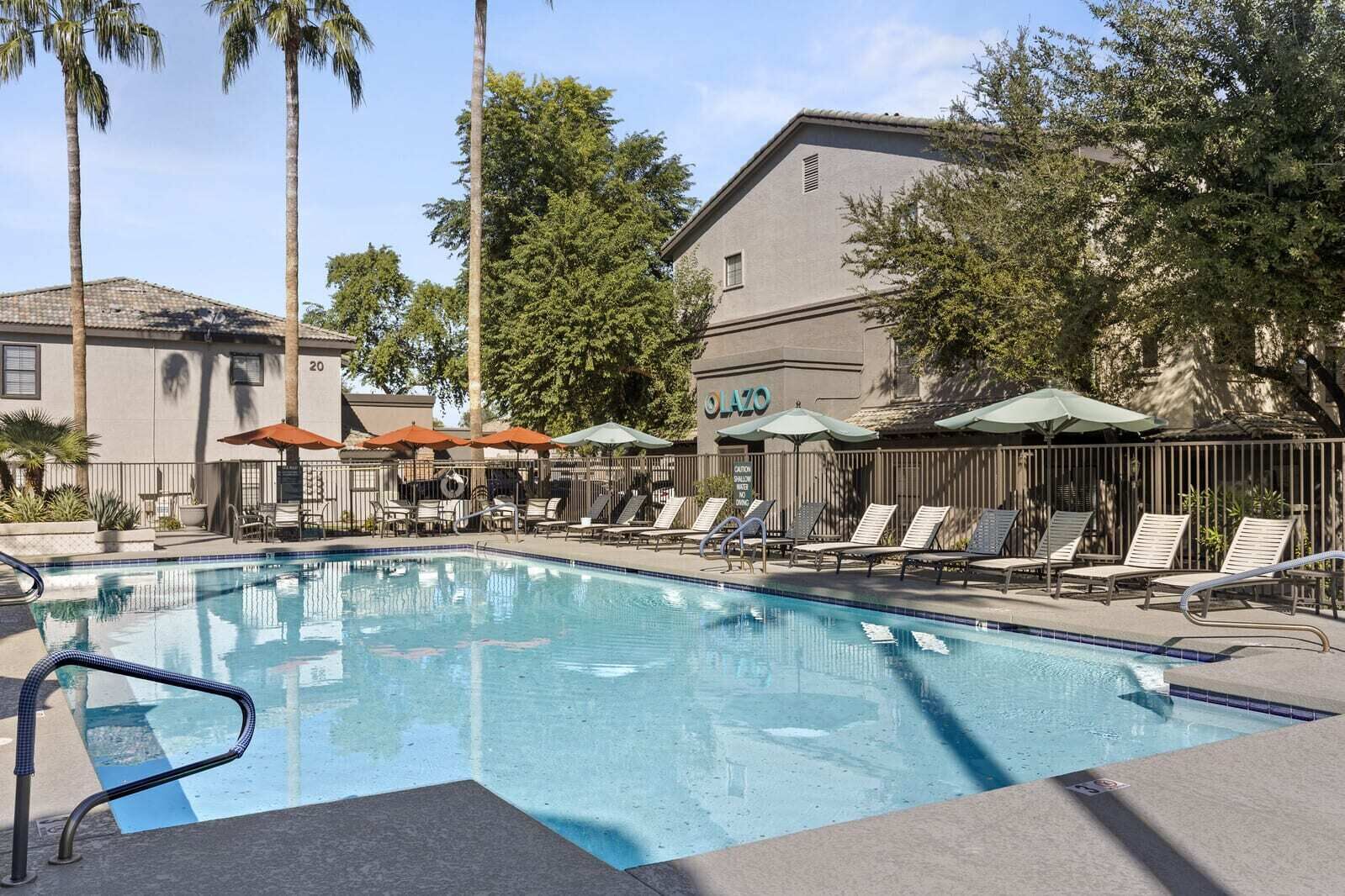 Ein Swimmingpool mit Liegestühlen und Sonnenschirmen, umgeben von Palmen und einem Gebäude mit der Aufschrift 'O'LAZO'.