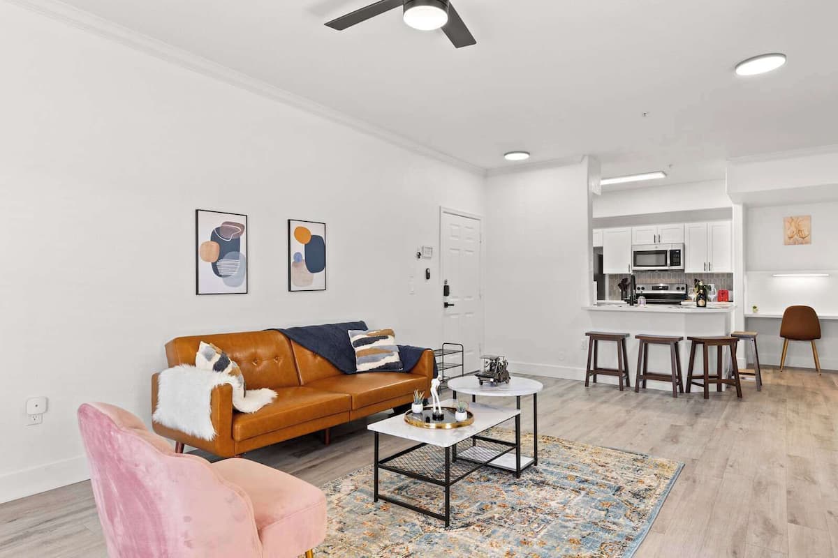 A modern living room with a brown leather sofa, a round coffee table, and a pink armchair. A kitchen with white cabinets and a bar area is visible in the background.