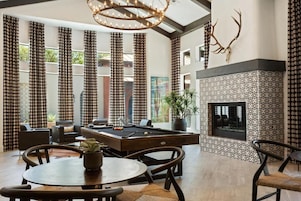 A modern living room with a pool table, comfortable seating, a fireplace, and a large window with curtains.