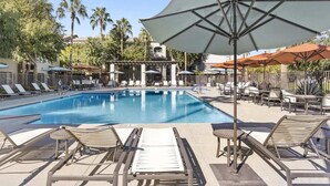 A pool area with lounge chairs and umbrellas, surrounded by palm trees and buildings. - CozySuites at Chandler (Chandler)