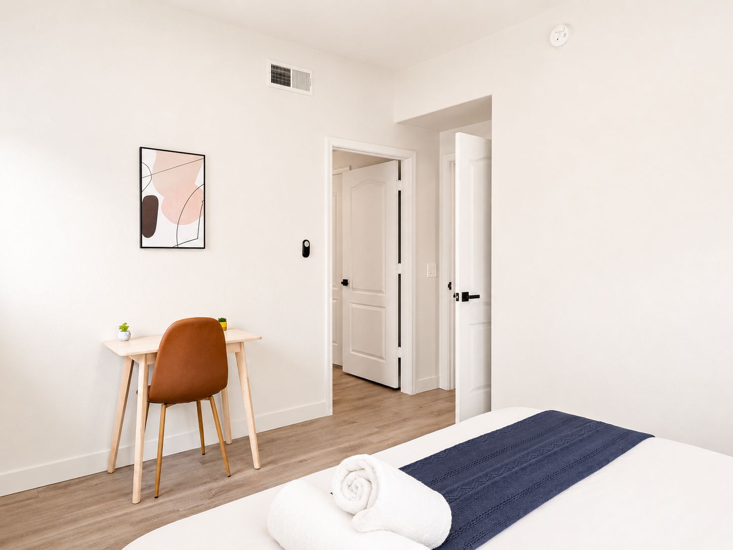 A hotel room with a bed, a desk with a chair, a door leading to another room, and a wall-mounted air vent.