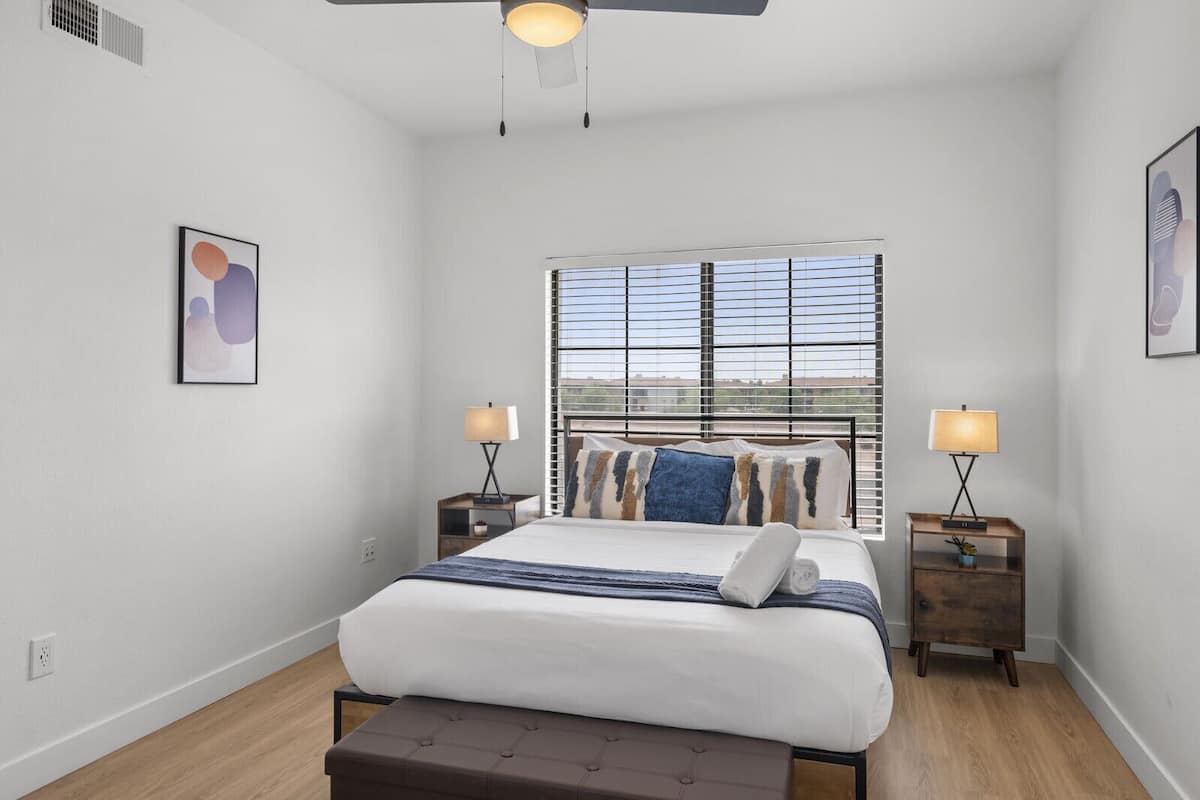 A bedroom with a large bed, two bedside tables with lamps, a ceiling fan, and a window with blinds.